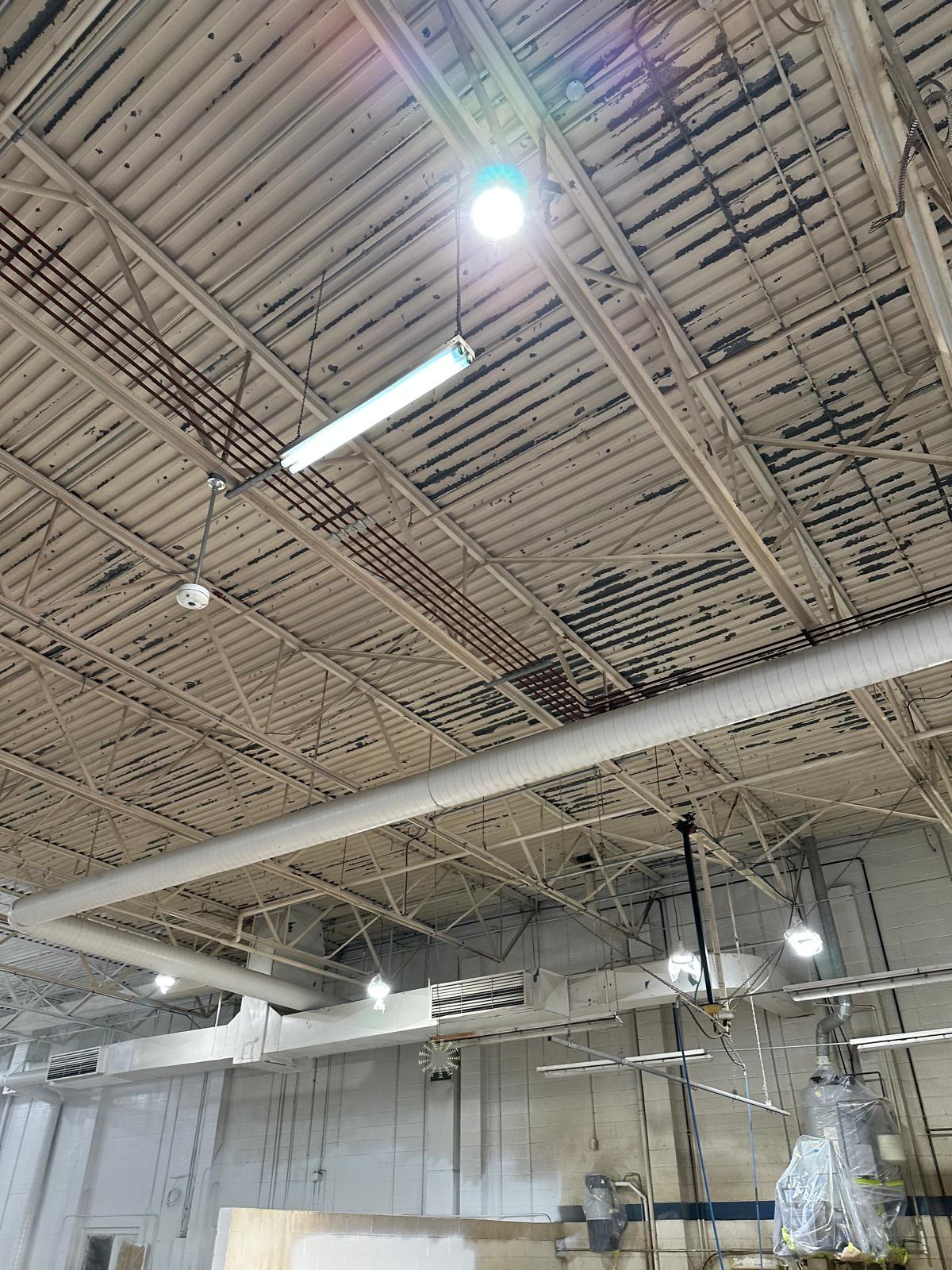 Shop ceiling before painting: steel roof deck peeling in long strips above the trusses and the round duct run